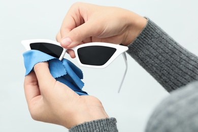 Photo of Woman wiping sunglasses with microfiber cleaning cloth on light background, closeup