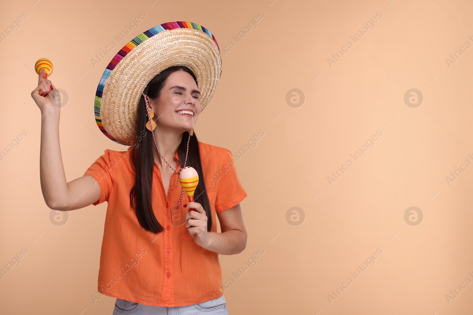 Photo of Young woman in Mexican sombrero hat dancing with maracas on beige background. Space for text