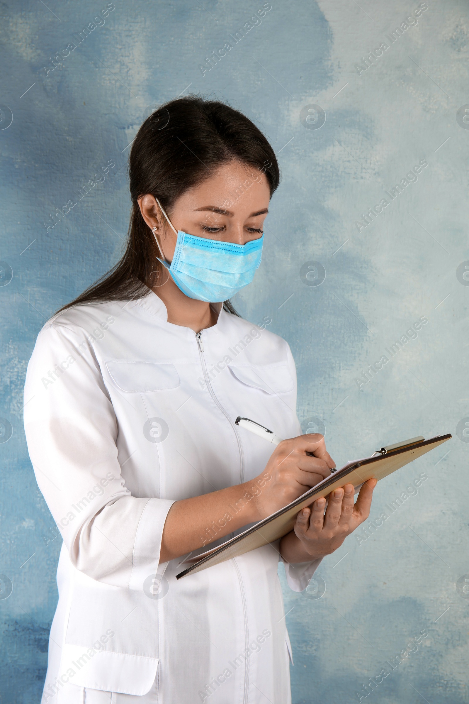 Photo of Doctor with disposable mask on face holding clipboard against light blue background