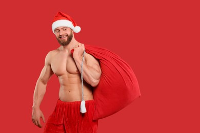 Photo of Muscular young man in Santa hat holding bag with presents on red background, space for text