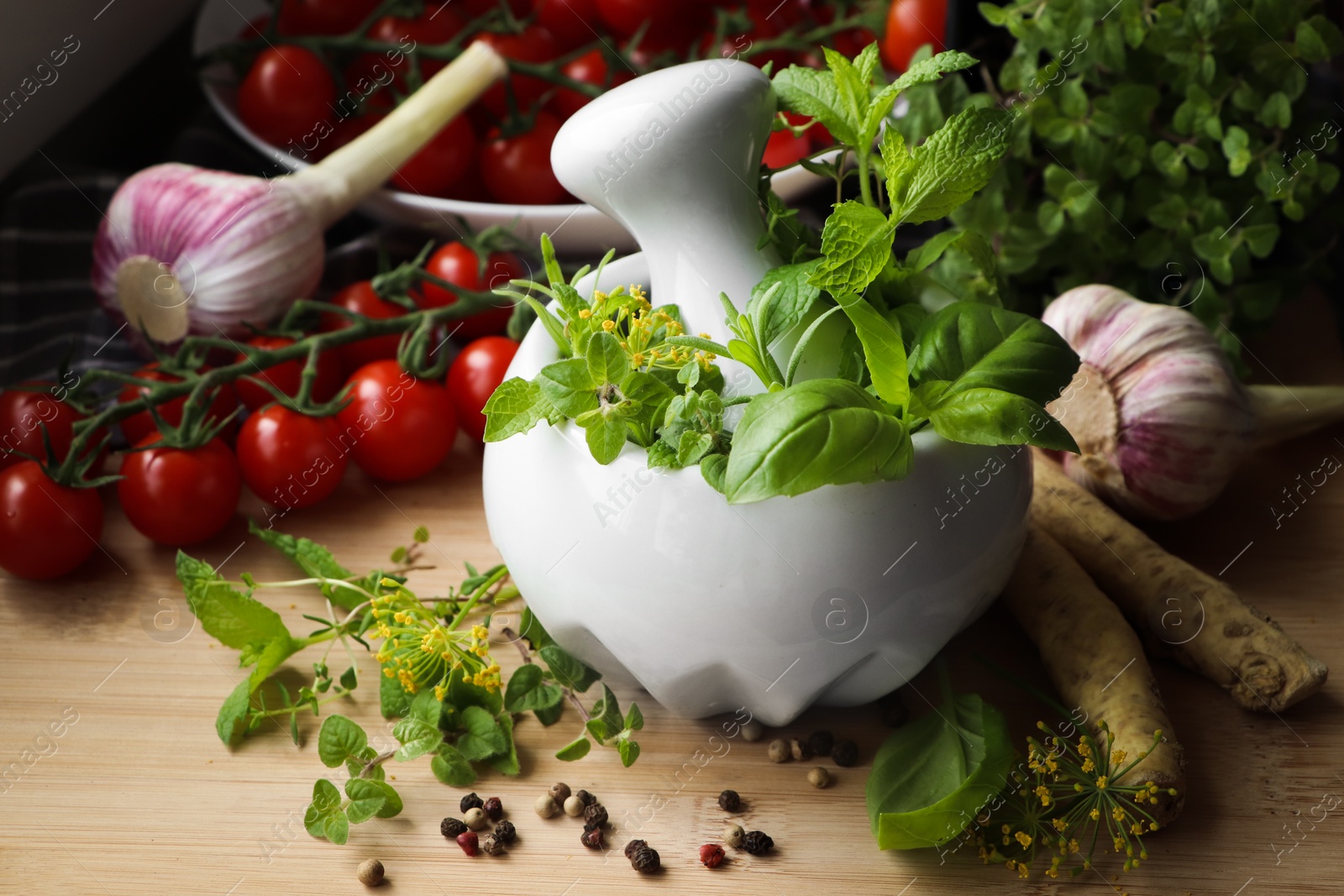 Photo of Mortar with fresh herbs near garlic, horseradish roots, black peppercorns and cherry tomatoes on wooden table, closeup