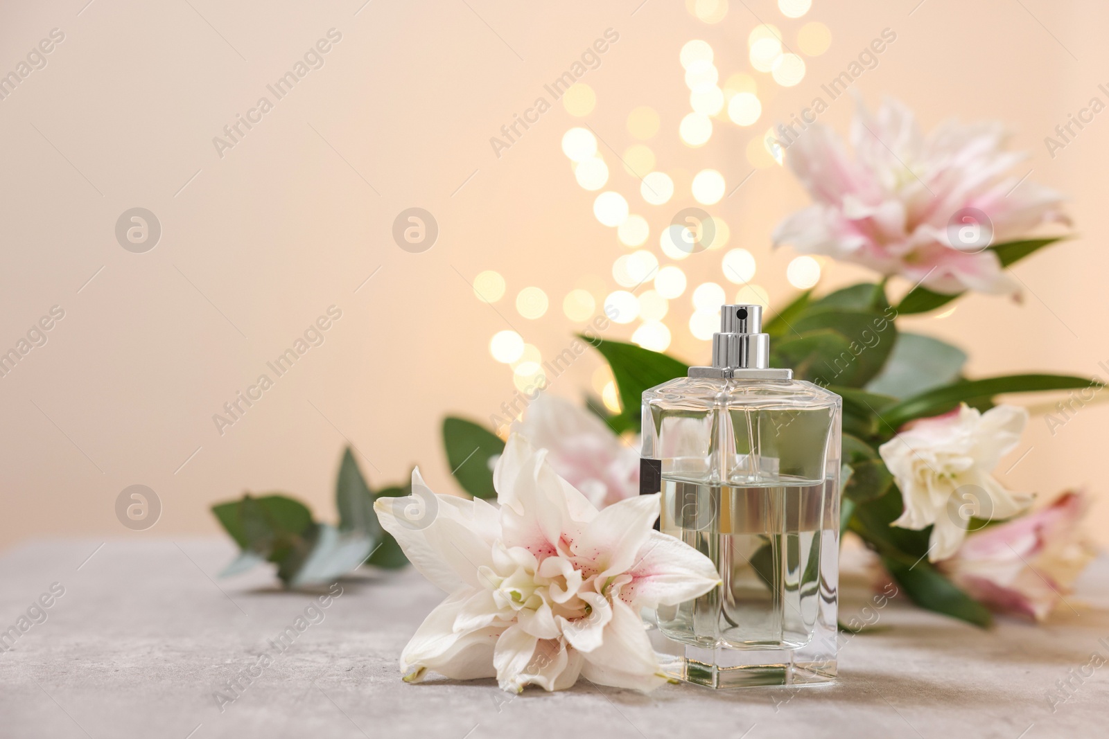 Photo of Bottle of perfume and beautiful lily flowers on table against beige background with blurred lights, space for text