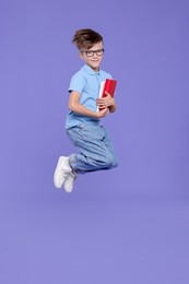 Photo of Cute schoolboy with books jumping on violet background, space for text