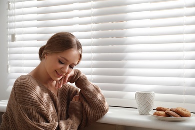 Beautiful young woman with cup of hot drink and cookies near window at home. Winter atmosphere