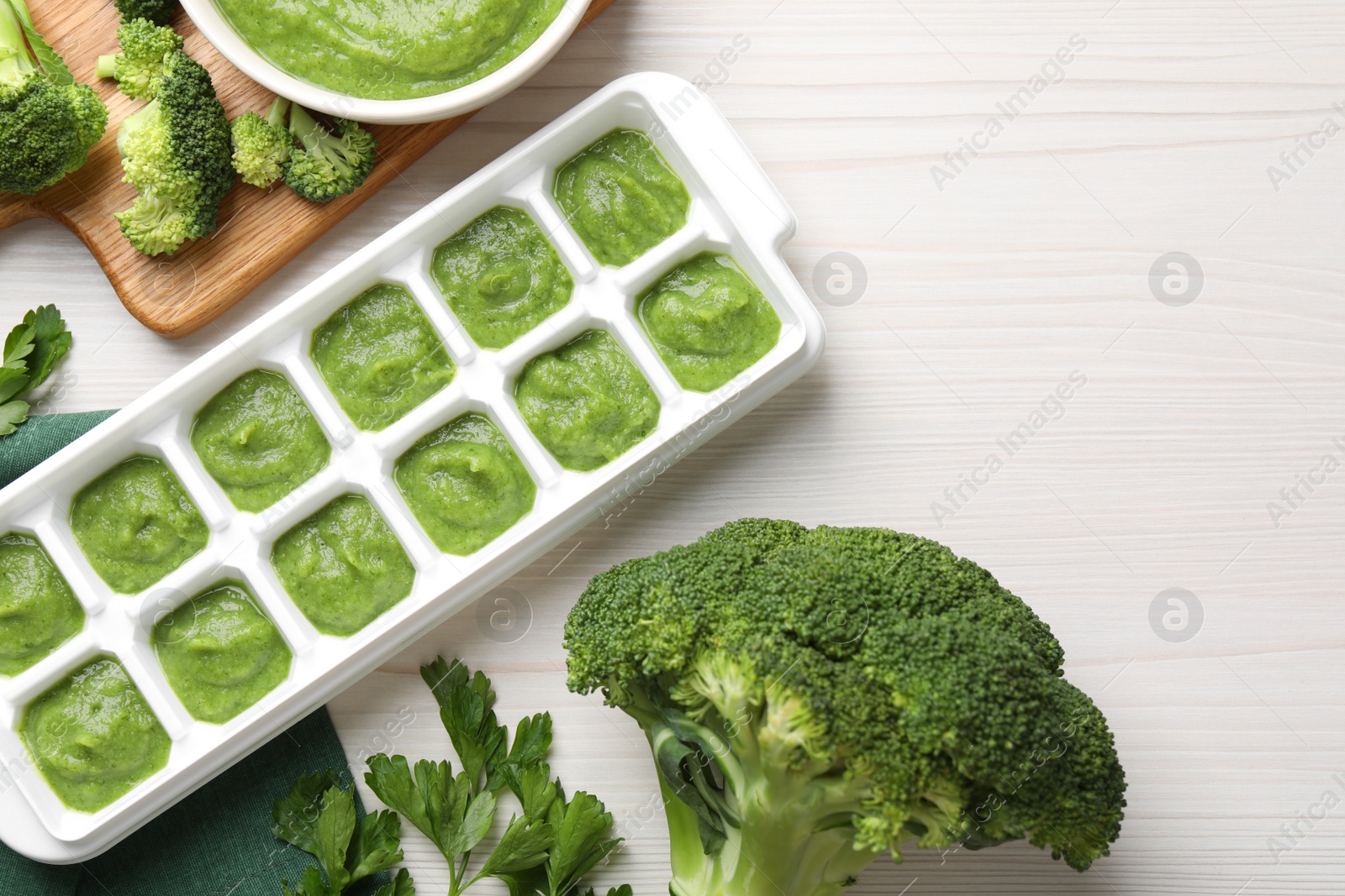 Photo of Broccoli puree in ice cube tray ready for freezing and ingredients on white wooden table, flat lay. Space for text