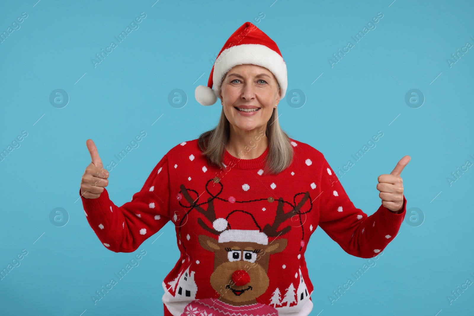 Photo of Happy senior woman in Christmas sweater and Santa hat showing thumbs up on light blue background