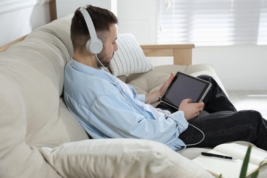 Photo of Young man with headphones using modern tablet for studying on sofa at home. Distance learning