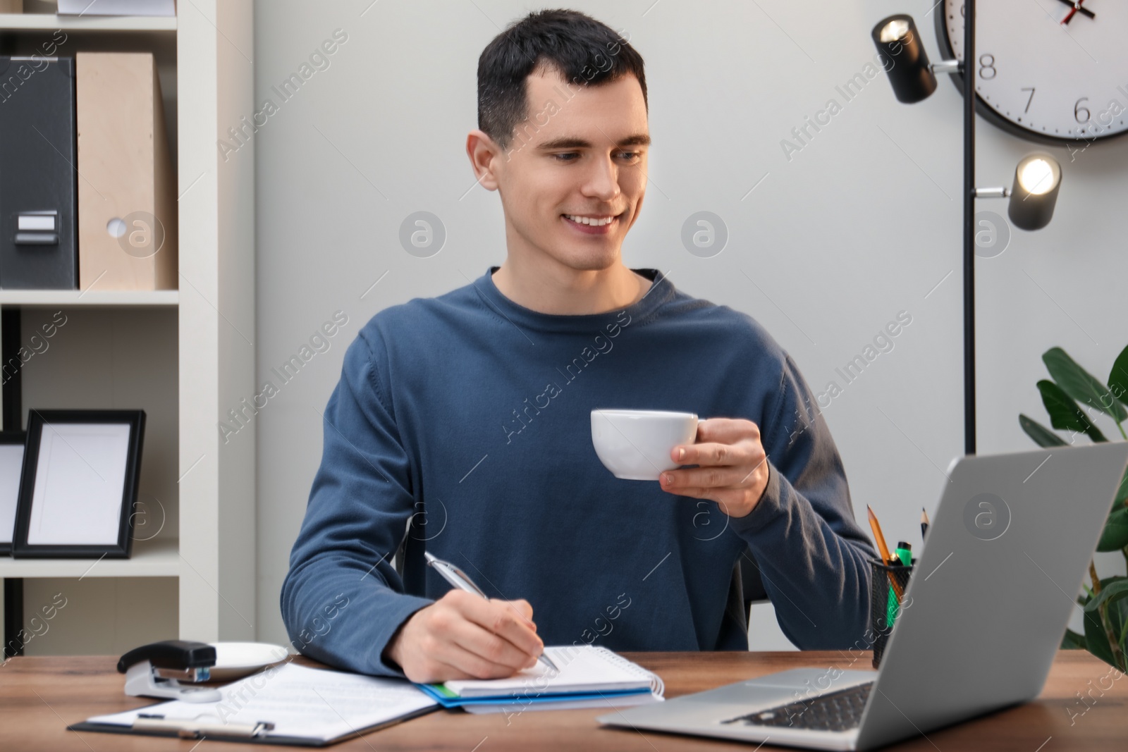 Photo of Man with cup of drink taking notes at wooden table in office