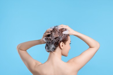 Young woman washing her hair with shampoo on light blue background, back view