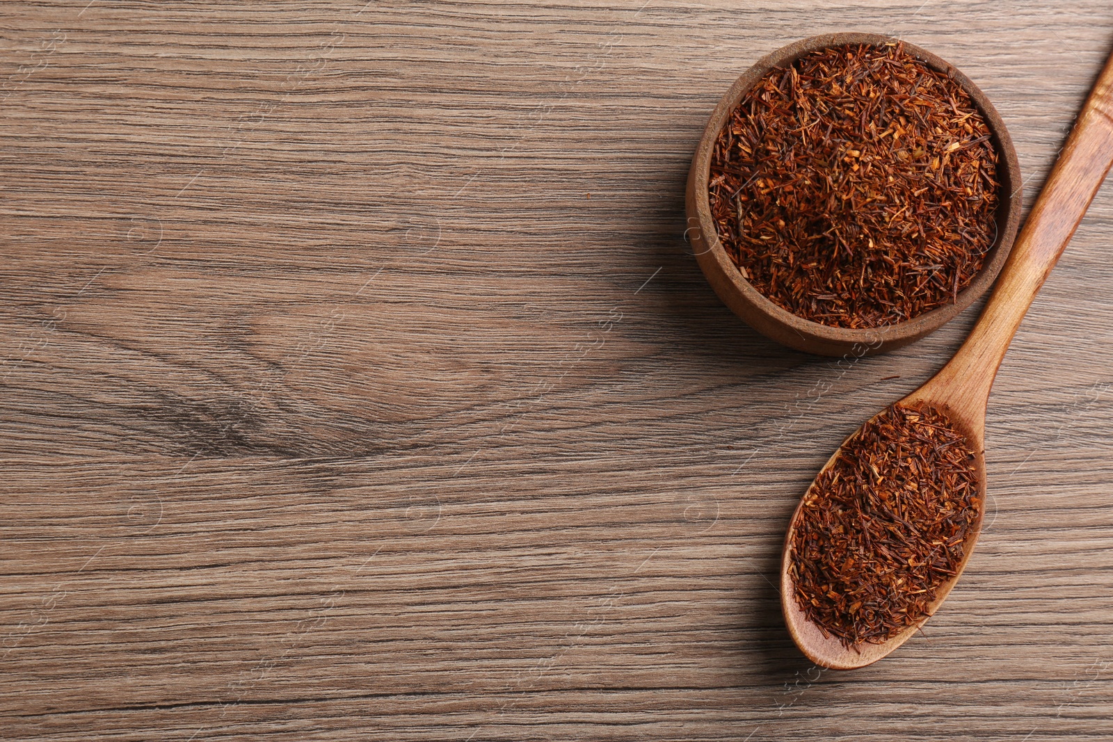 Photo of Spoon and bowl with dry rooibos leaves on wooden table, flat lay. Space for text 