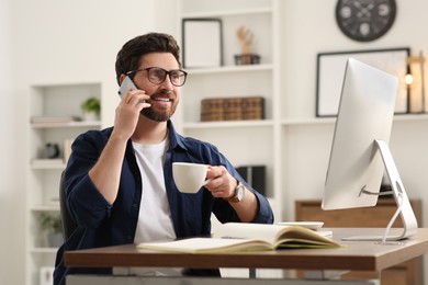 Home workplace. Happy man with cup of drink talking on smartphone while working with computer at wooden desk in room