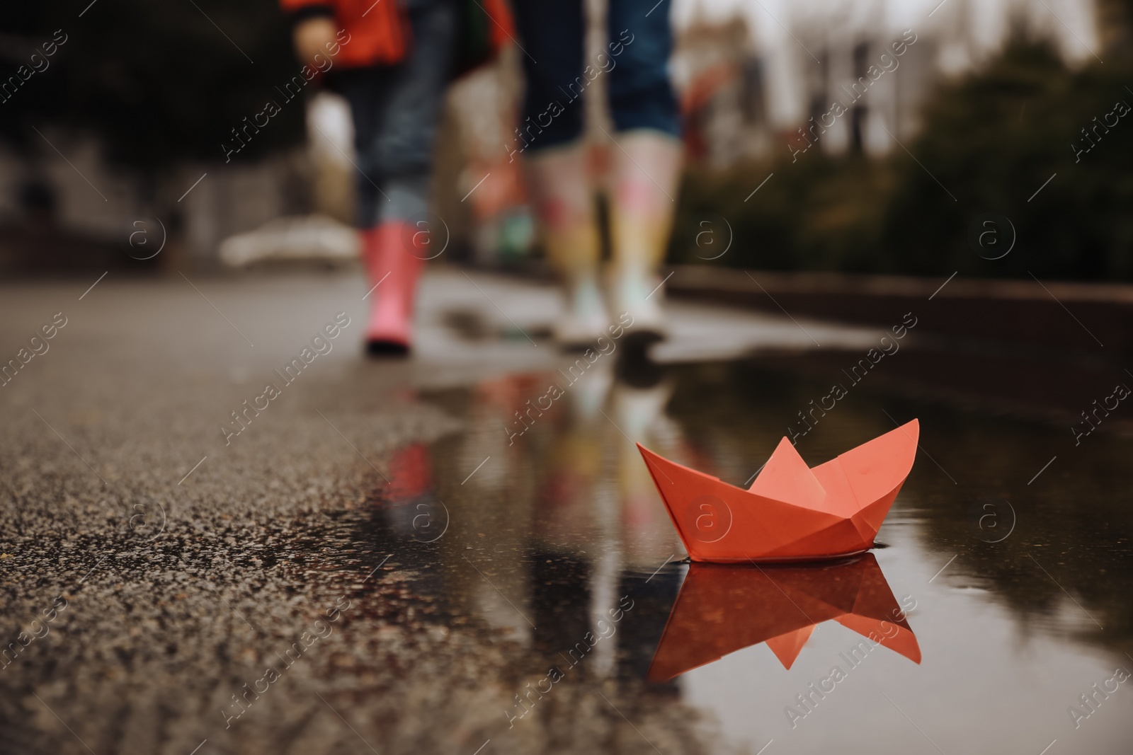 Photo of Little girl with her mother walking outdoors, focus on  paper boat in puddle