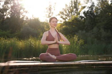 Photo of Beautiful woman practicing Padmasana on yoga mat on wooden pier near pond. Lotus pose