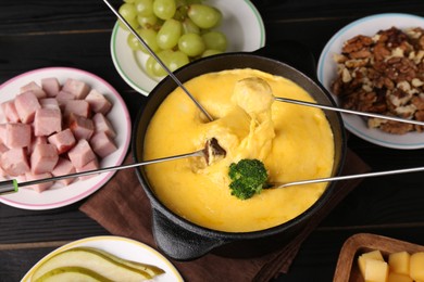 Photo of Dipping different products into fondue pot with melted cheese on black wooden table, above view