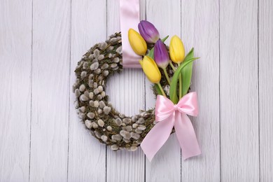 Photo of Wreath made of beautiful willow, colorful tulip flowers and pink bow hanging on white wooden background