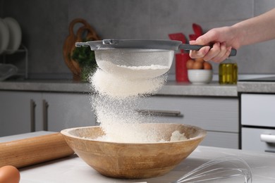 Photo of Woman sieving flour into bowl at white wooden table in kitchen, closeup