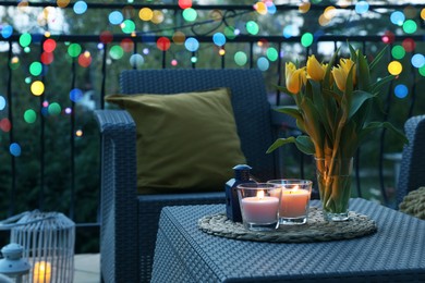 Soft pillows, blanket, burning candles and yellow tulips on rattan garden furniture in evening