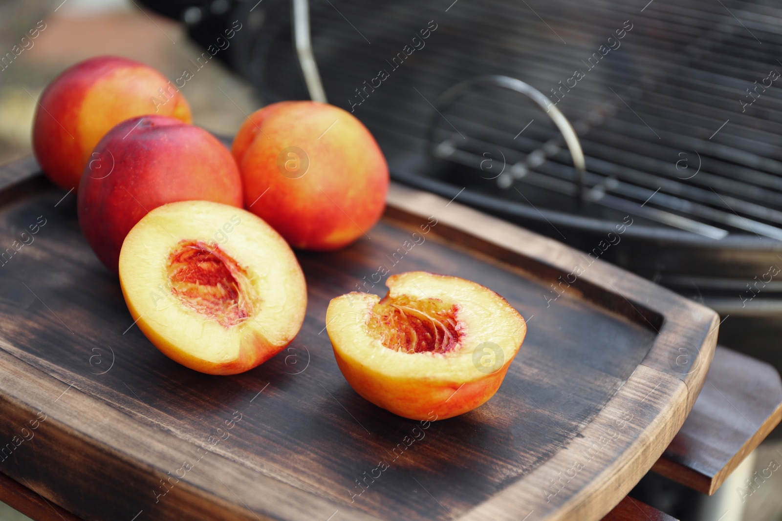 Photo of Fresh cut and whole peaches on wooden board near modern grill outdoors, closeup