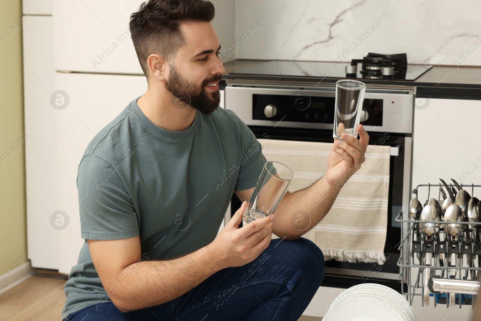 Photo of Smiling man loading dishwasher with glasses in kitchen