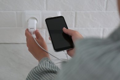 Woman with mobile phone plugging charger in socket on white brick wall, closeup