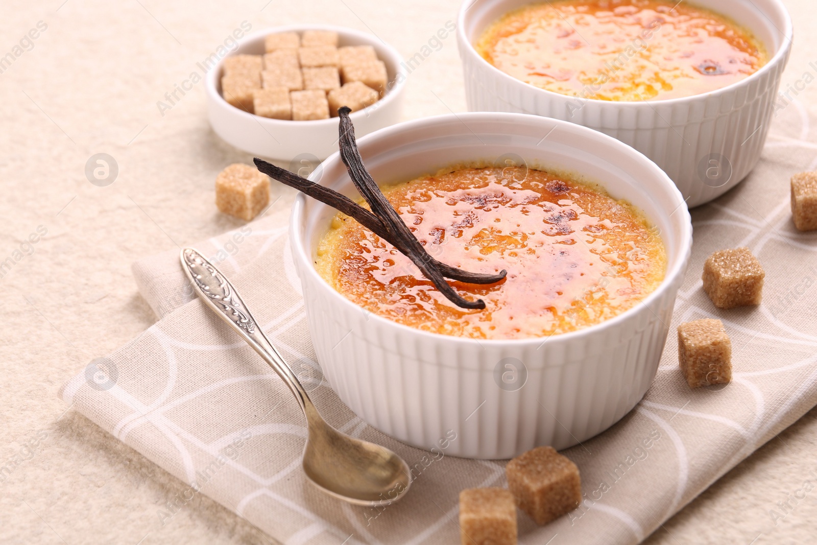 Photo of Delicious creme brulee in bowl, vanilla pods, sugar cubes and spoon on light textured table, closeup
