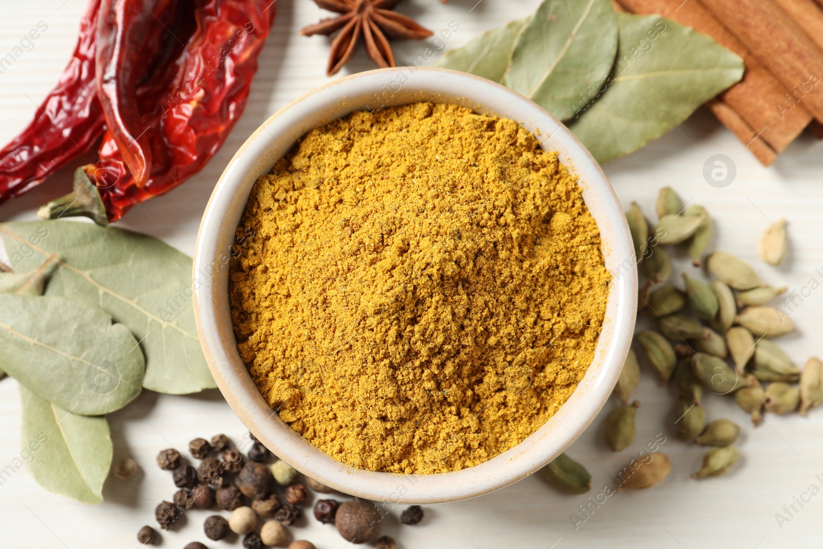 Photo of Curry powder in bowl and other spices on white wooden table, flat lay