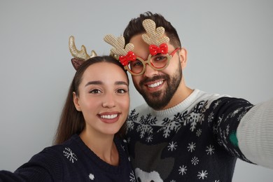 Photo of Happy young couple in Christmas sweaters, reindeer headband and funny glasses taking selfie on grey background