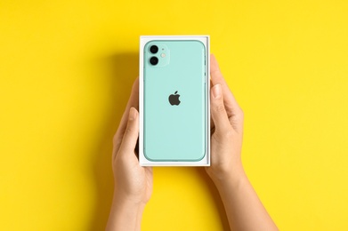 Photo of MYKOLAIV, UKRAINE - JULY 10, 2020: Woman holding box with new modern Iphone 11 Green on yellow background, top view