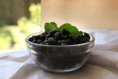 Bowl of delicious ripe black mulberries with green leaves on white fabric indoors