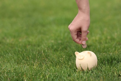 Photo of Young woman putting coin into piggy bank on green grass outdoors, closeup. Space for text