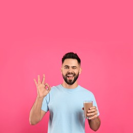 Young man with glass of chocolate milk showing Ok on pink background