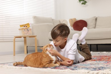 Photo of Little boy petting cute ginger cat on carpet at home