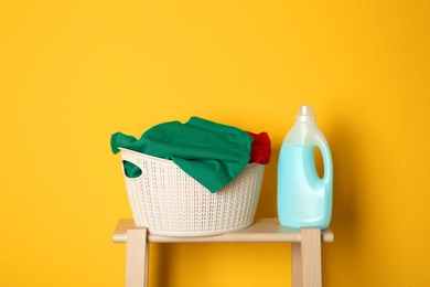 Photo of Laundry basket with dirty clothes and detergent on wooden stool against yellow background