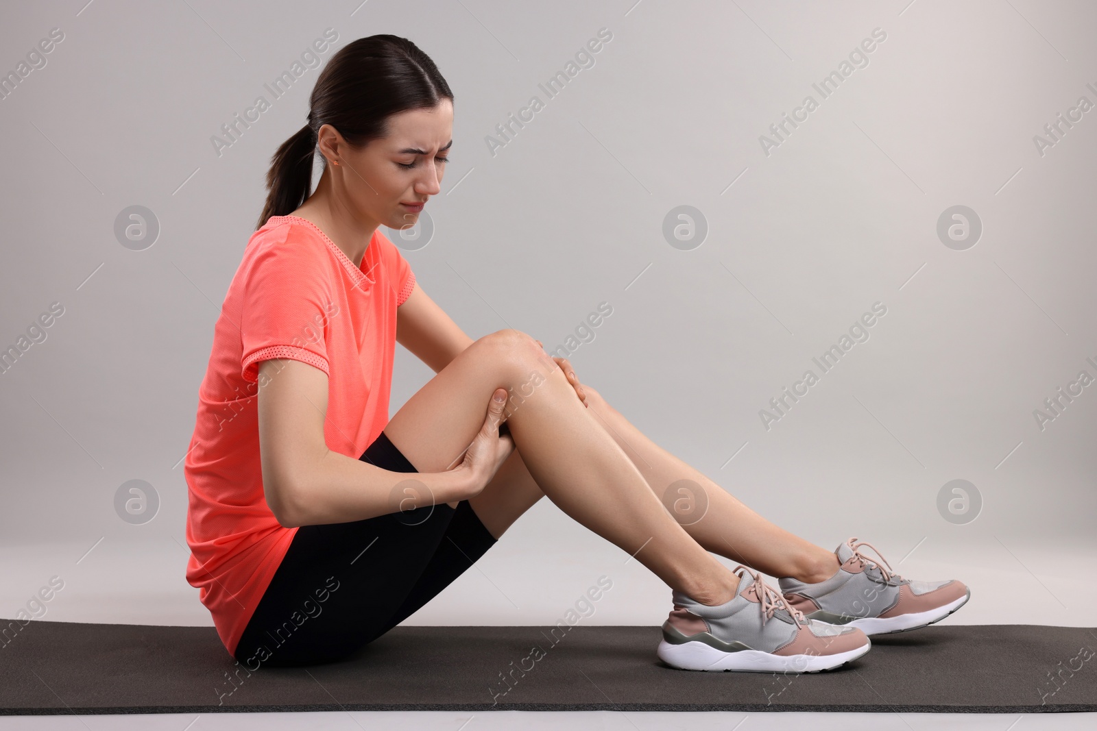 Photo of Young woman suffering from leg pain on exercise mat against grey background