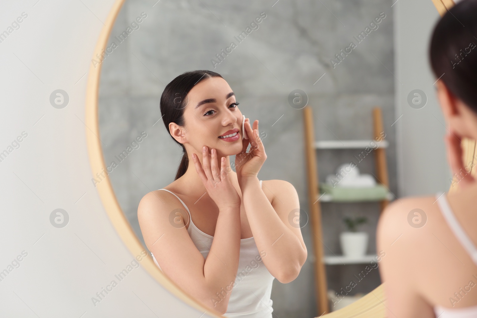 Photo of Beautiful woman removing makeup with cotton pad near mirror indoors