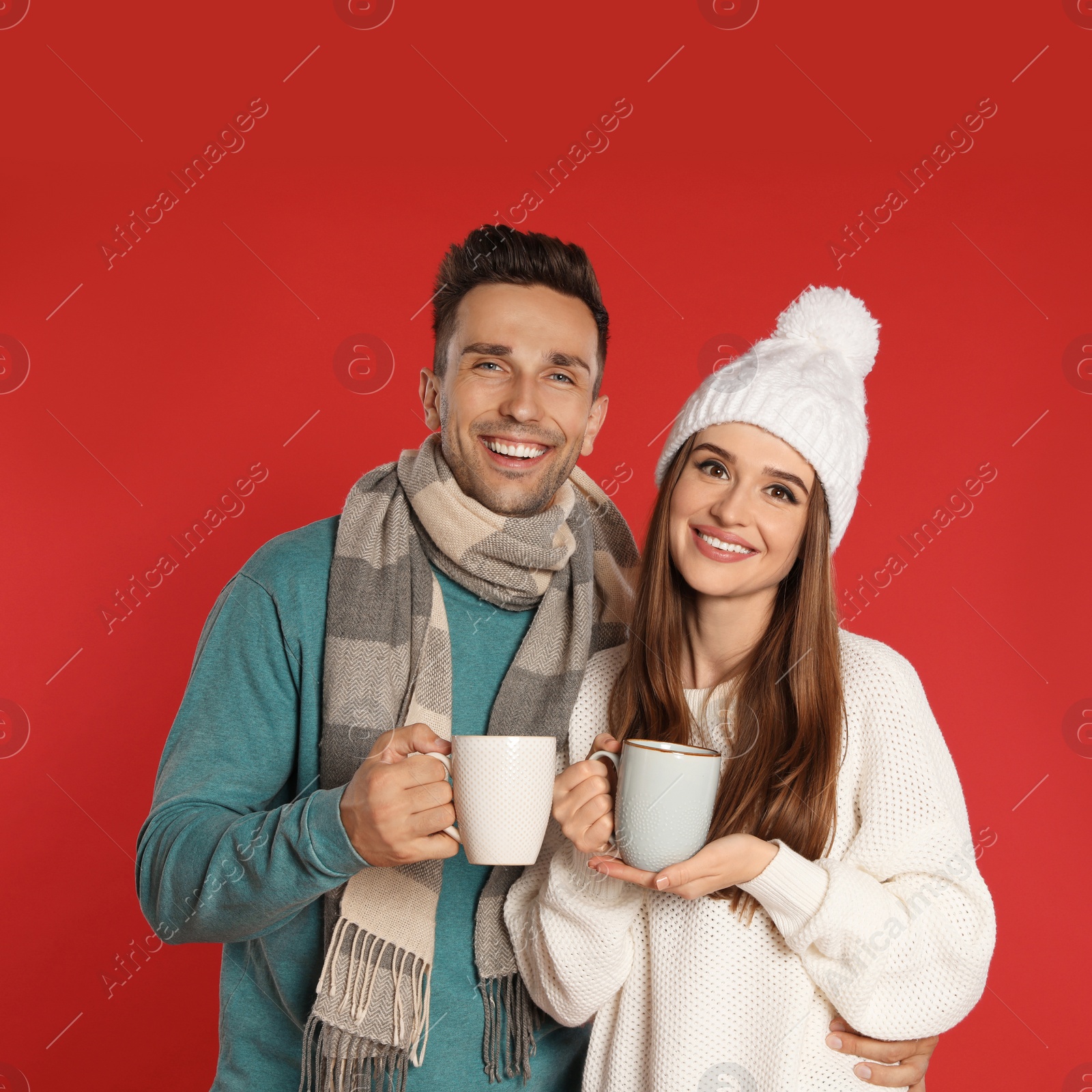 Photo of Happy young couple in warm clothes with cups on red background. Winter season