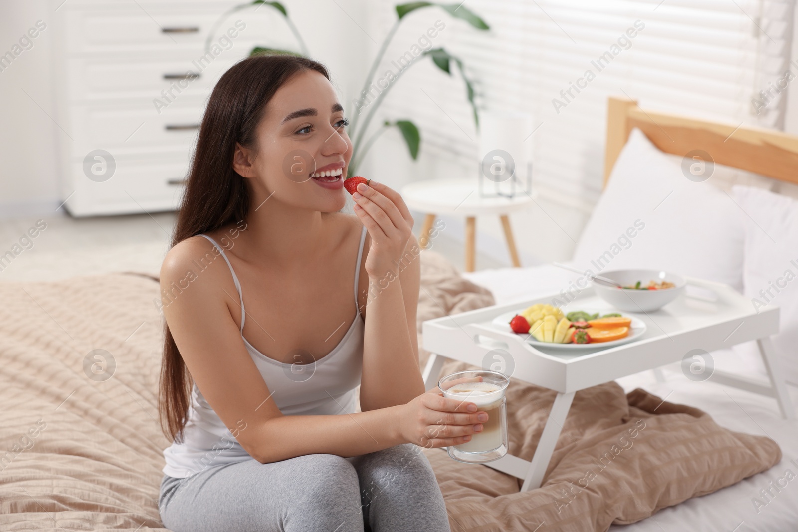 Photo of Happy young woman having breakfast near white tray on bed at home