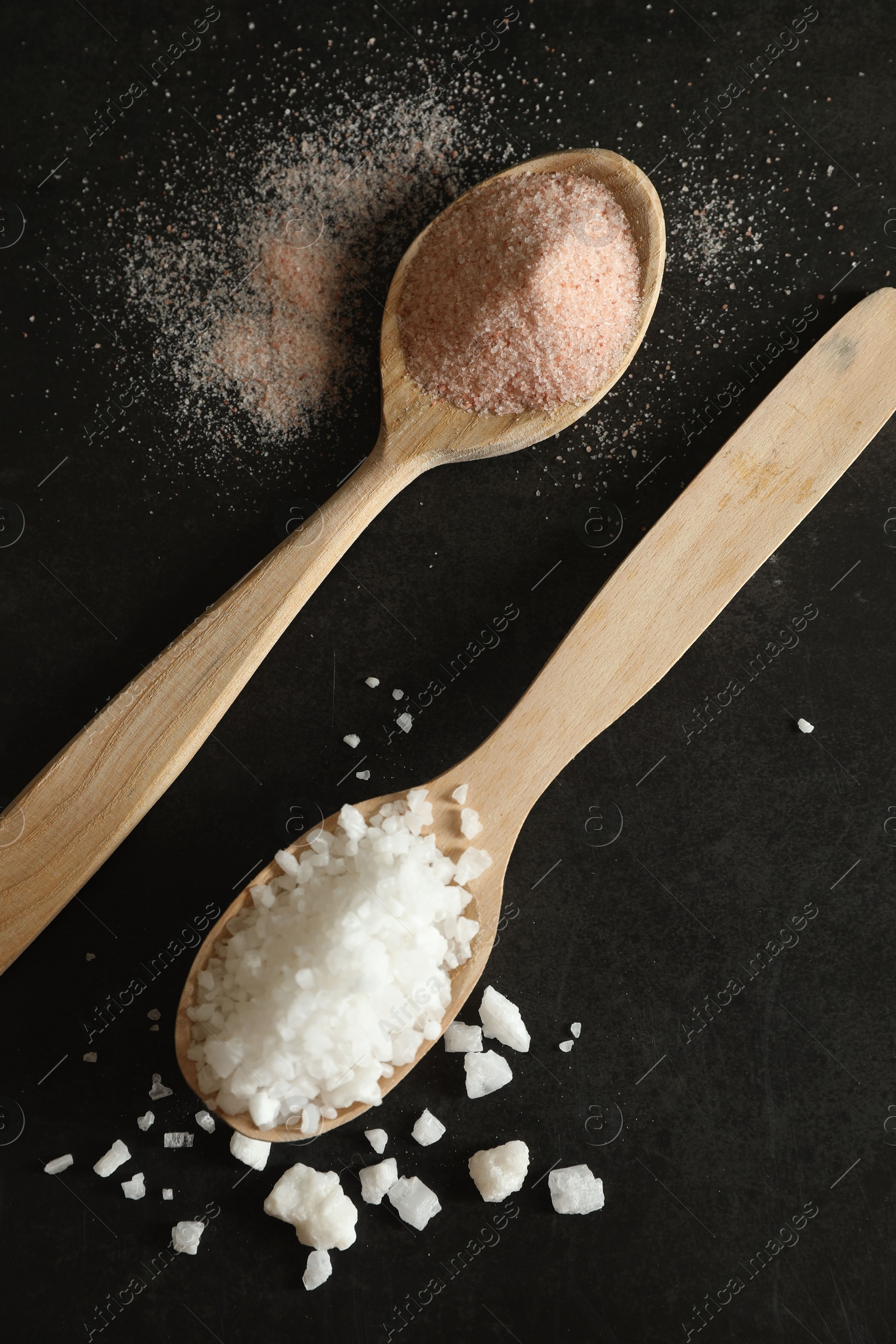 Photo of Different types of organic salt in spoons on black table, flat lay