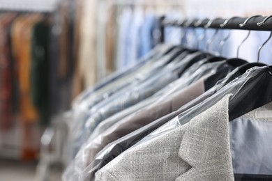 Photo of Dry-cleaning service. Many different clothes in plastic bags hanging on rack indoors, closeup