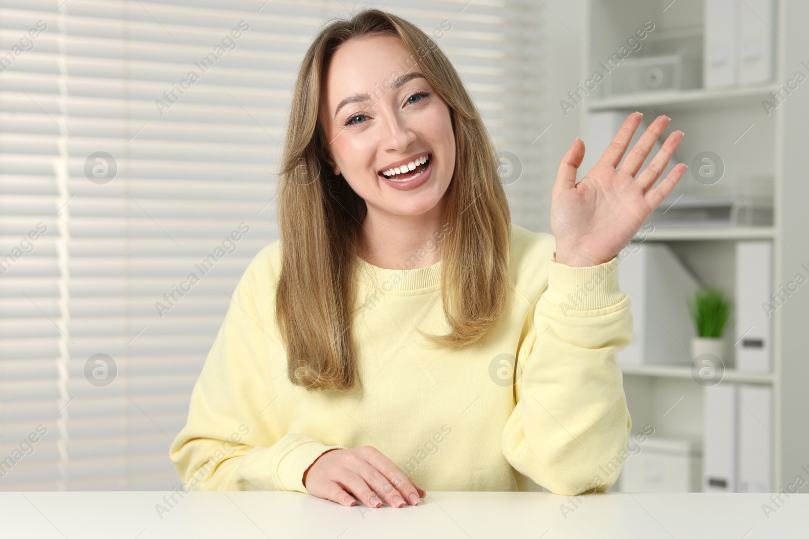 Photo of Young woman waving hello during video chat at white table indoors, view from webcam