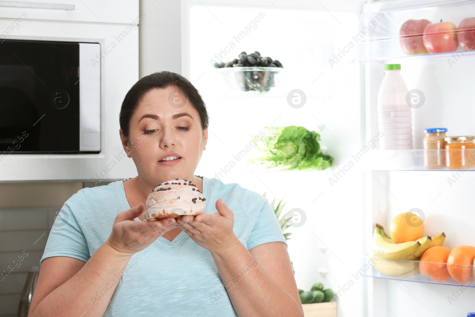 Photo of Woman holding tasty dessert near fridge in kitchen. Diet failure