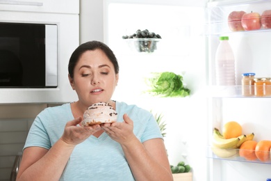 Woman holding tasty dessert near fridge in kitchen. Diet failure