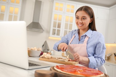 Photo of Happy woman cutting mushroom while watching online cooking course via laptop in kitchen