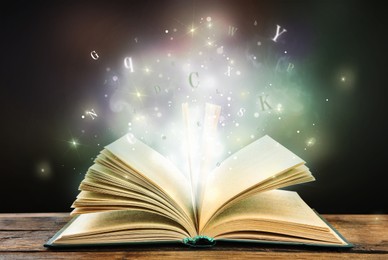 Open book with magic light and glowing letters flying out of it on wooden table against black background