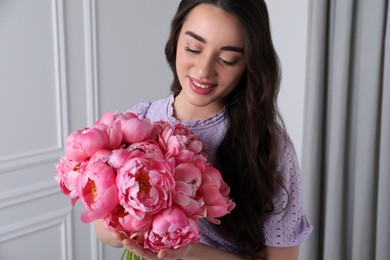 Photo of Beautiful young woman with bouquet of pink peonies indoors
