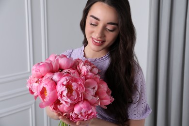 Beautiful young woman with bouquet of pink peonies indoors