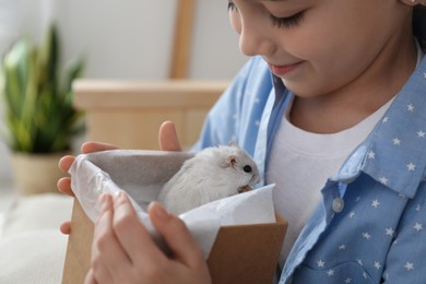 Happy little girl holding gift box with cute hamster at home