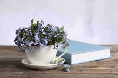 Beautiful forget-me-not flowers in cup, saucer and book on wooden table against light background, closeup