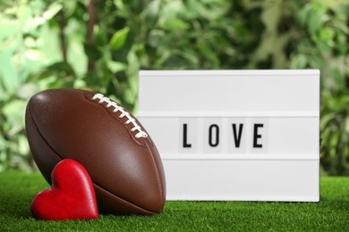 Photo of American football ball, lightbox with word Love and heart on green grass against blurred background