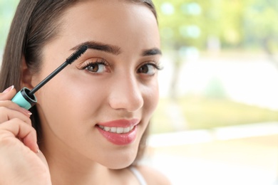 Attractive young woman applying mascara on her eyelashes against blurred background